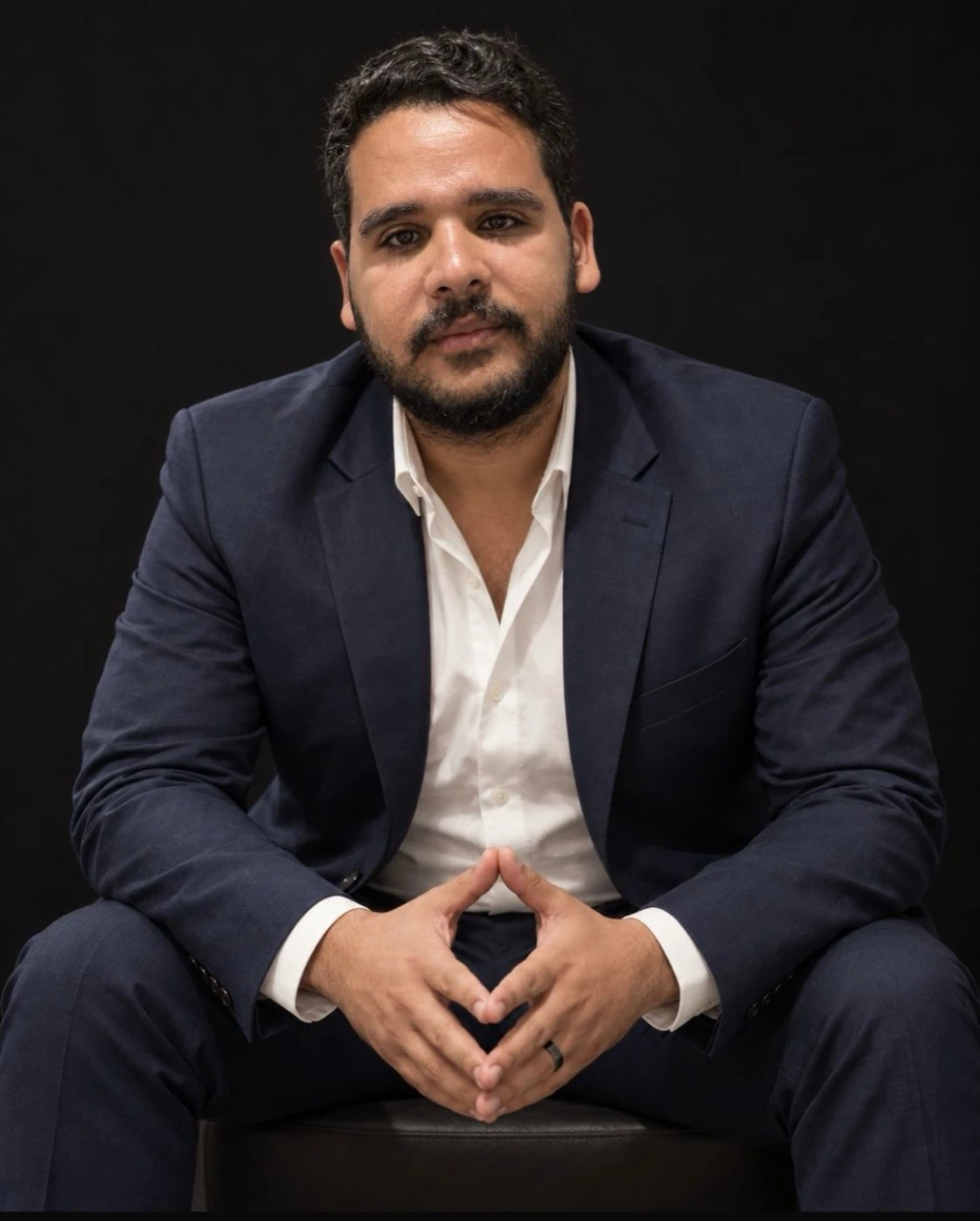 Ali Ashraf in a navy suit against a dark studio background.